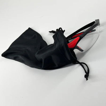 A pair of Karakal Adult Pro 3000 Sports Eye Protection goggles by Karakal, featuring clear anti-fog polycarbonate lenses and black-and-red frames, is partially pulled from a black drawstring pouch on a white surface.