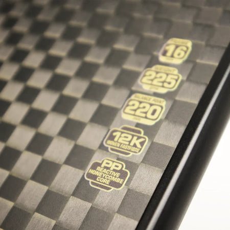 Close-up of the Karakal Pure Touch Gold Pickleball Paddle by Karakal, featuring a black and gray checkered design, gold icons, and specs: 16, 225, 220, power core 12K woven fastfire, with a textured PP reactive honeycomb core.
