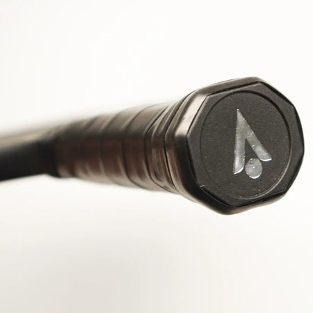 Close-up of the black, textured handlebar grip on the Karakal Pure Touch Gold Pickleball Paddle, featuring a hexagonal black cap with Karakal's stylized A logo and dot. The grip stands out boldly against a soft, white background.