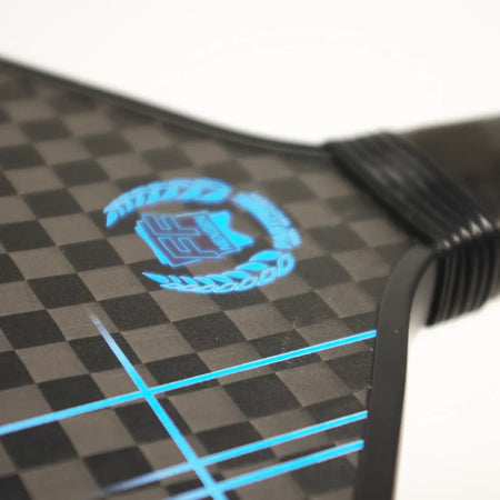 Close-up of the Karakal Pure Touch Blue Pickleball Paddle by Karakal, featuring a black and gray carbon fiber textured surface with blue lines, a blue logo with laurel wreath design, and a partially visible black handle.