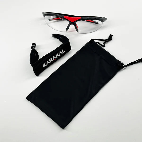 Karakal Adult Pro 3000 Sports Eye Protection by Karakal: clear safety glasses with black and red accents, anti-fog polycarbonate eye guards, adjustable black strap, and drawstring pouch. Displayed on a white background.