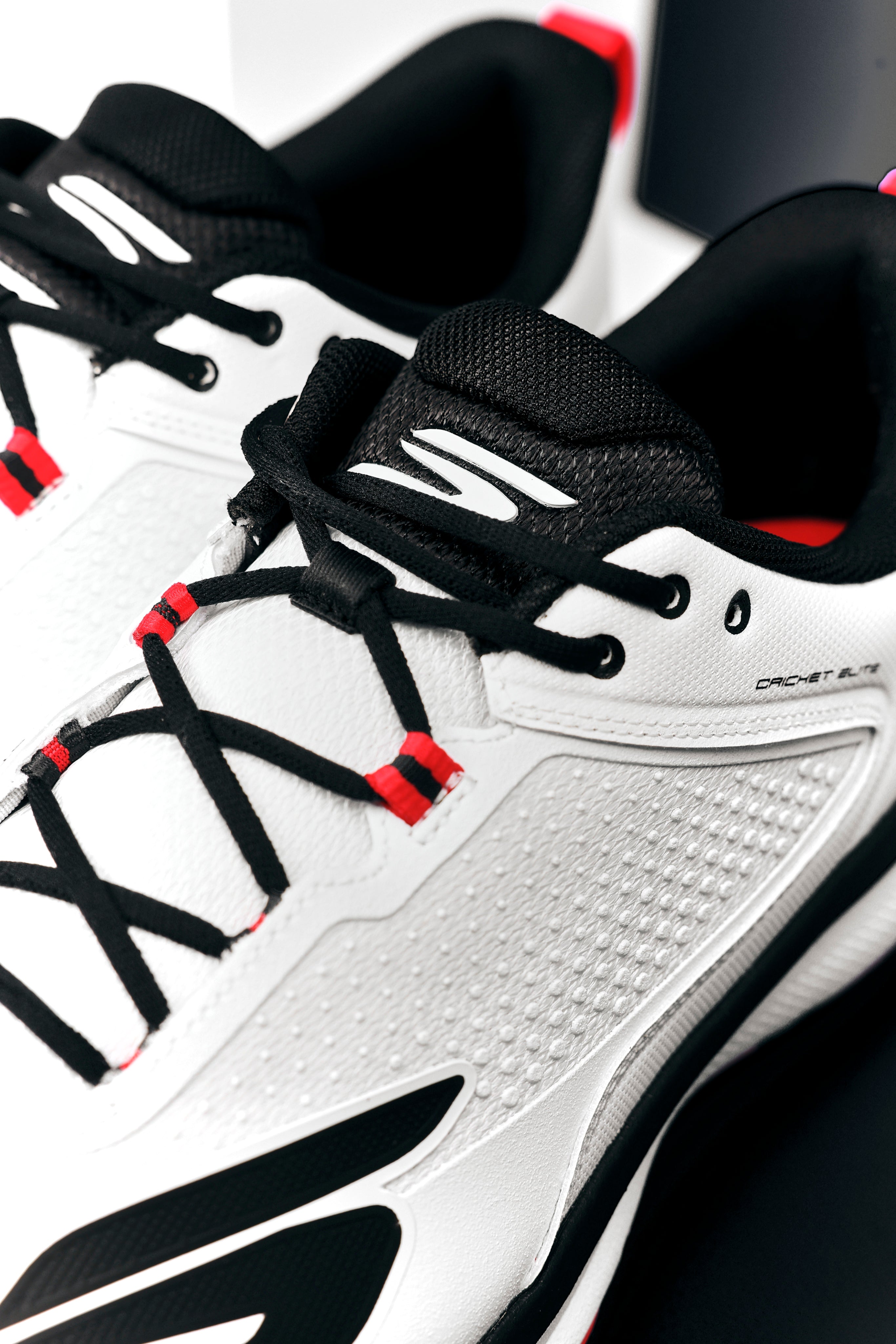 Close-up of Skechers Cricket Elite Cricket Shoe, white with black laces, black and red accents, and textured side panels. Designed by Skechers for superior grip on the field.