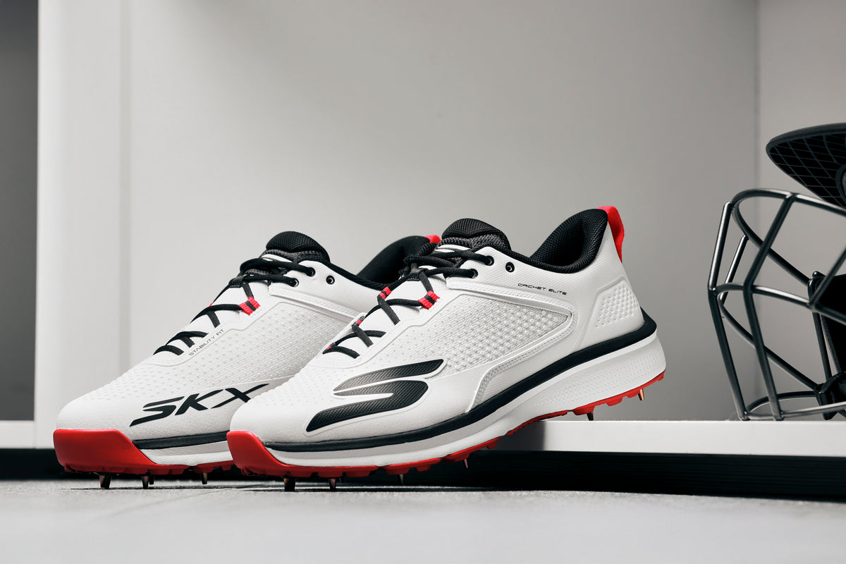 A pair of Skechers Cricket Elite Cricket Shoes in white with black and red accents, SKX logo, and Arch Fit insole, placed side by side on a white surface in a modern minimalistic room.
