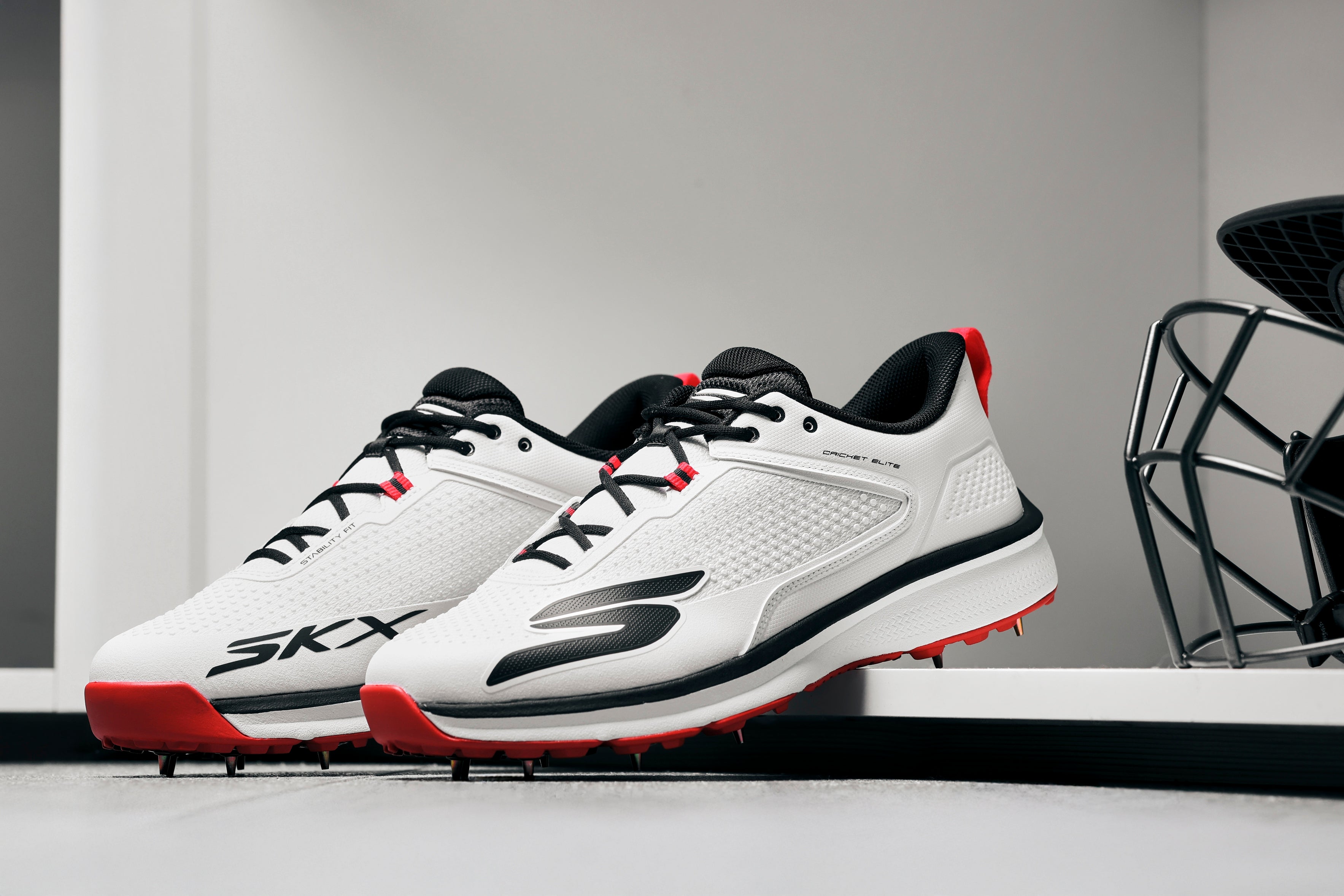 A pair of Skechers Cricket Elite Cricket Shoes in white with black and red accents, SKX logo, and Arch Fit insole, placed side by side on a white surface in a modern minimalistic room.