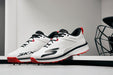 A pair of Skechers Cricket Elite Cricket Shoes in white with black and red accents, SKX logo, and Arch Fit insole, placed side by side on a white surface in a modern minimalistic room.