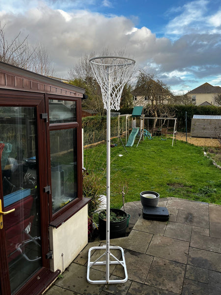 The Gilbert Academy Netball Post V2 by Gilbert stands on a stone patio near a conservatory, overlooking a grassy backyard with a swing set, potted plants, and garden shed under a partly cloudy sky.