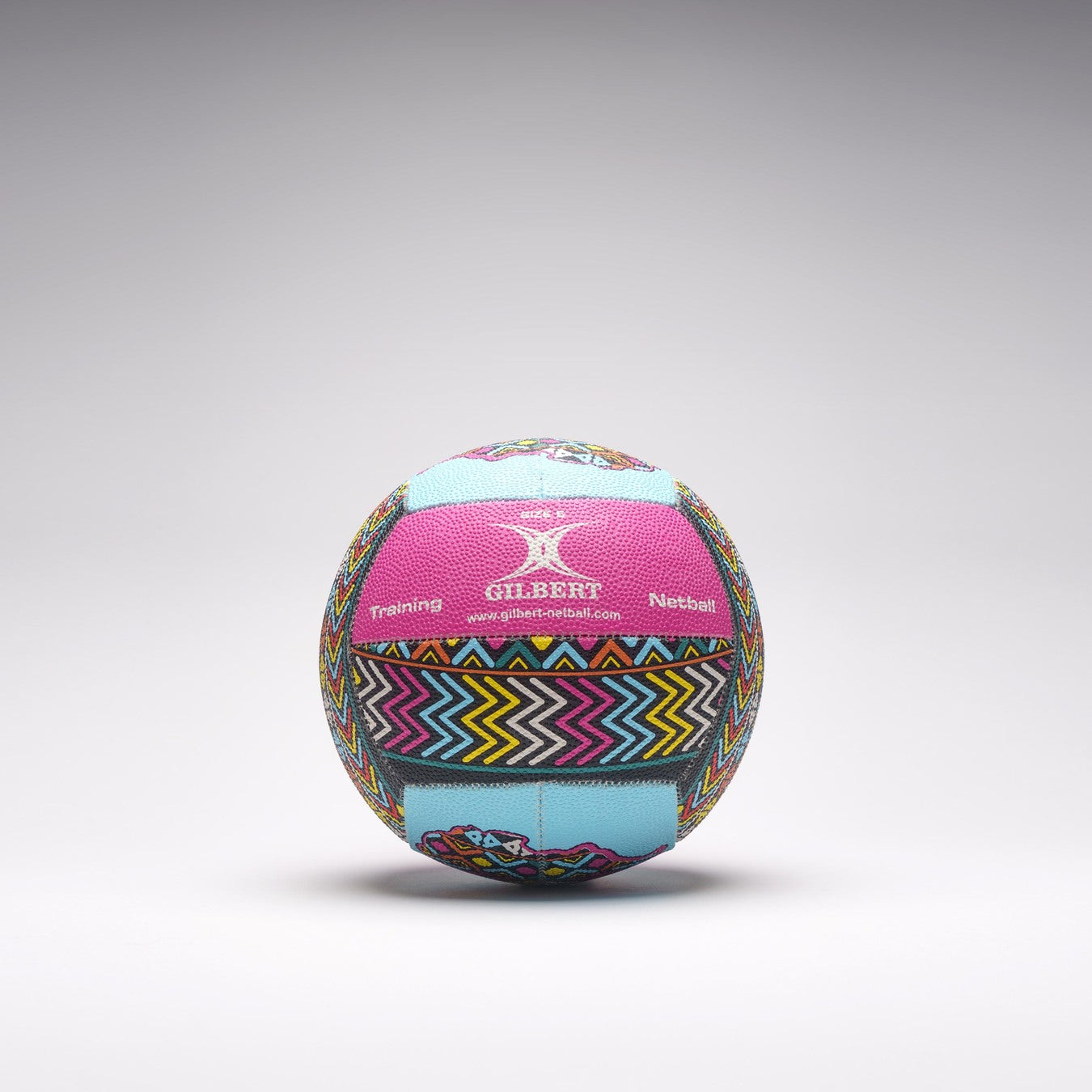 A vibrant Gilbert Ambassador Signature Netball in size 5, featuring zigzag and geometric pink, blue, yellow, and black patterns, rests on a light gray surface. Both "Gilbert" and "Training" are visible on this signature ball.