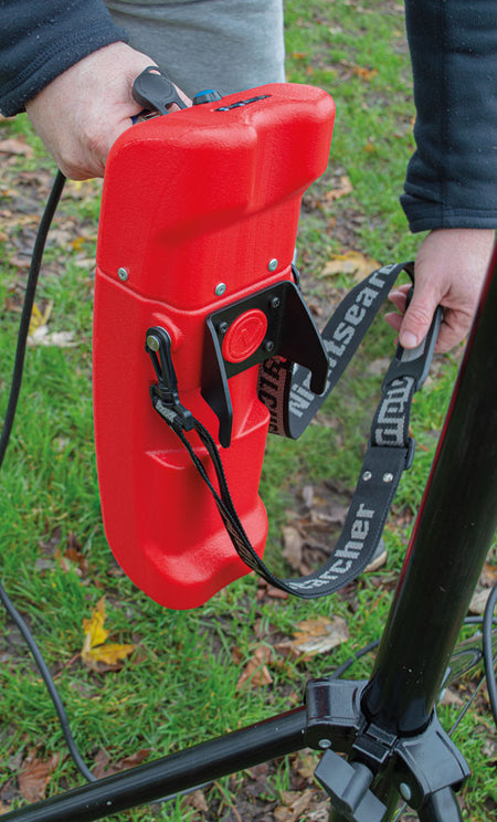 A person attaches a Nightsearcher SportStar Portable LED Floodlight with a black strap to a metal pole outdoors on green grass.