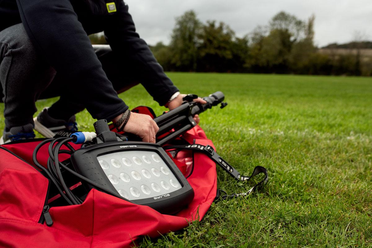 A person kneels on grass assembling gear from a red bag. Shown are a Nightsearcher SportStar Portable LED Floodlight and a tripod, with trees and a cloudy sky visible in the background.