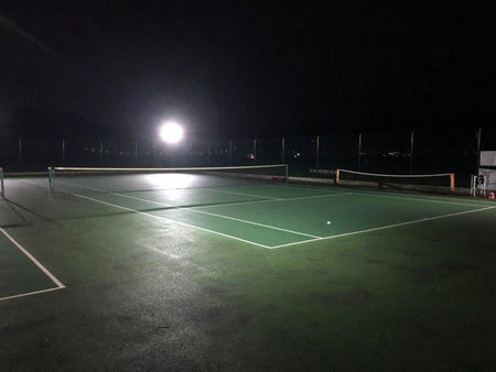 A tennis court is illuminated at night by the Nightsearcher SportStar Portable LED Floodlight (20,000 lumens), casting shadows and highlighting the green court surface and white lines against a dark background.