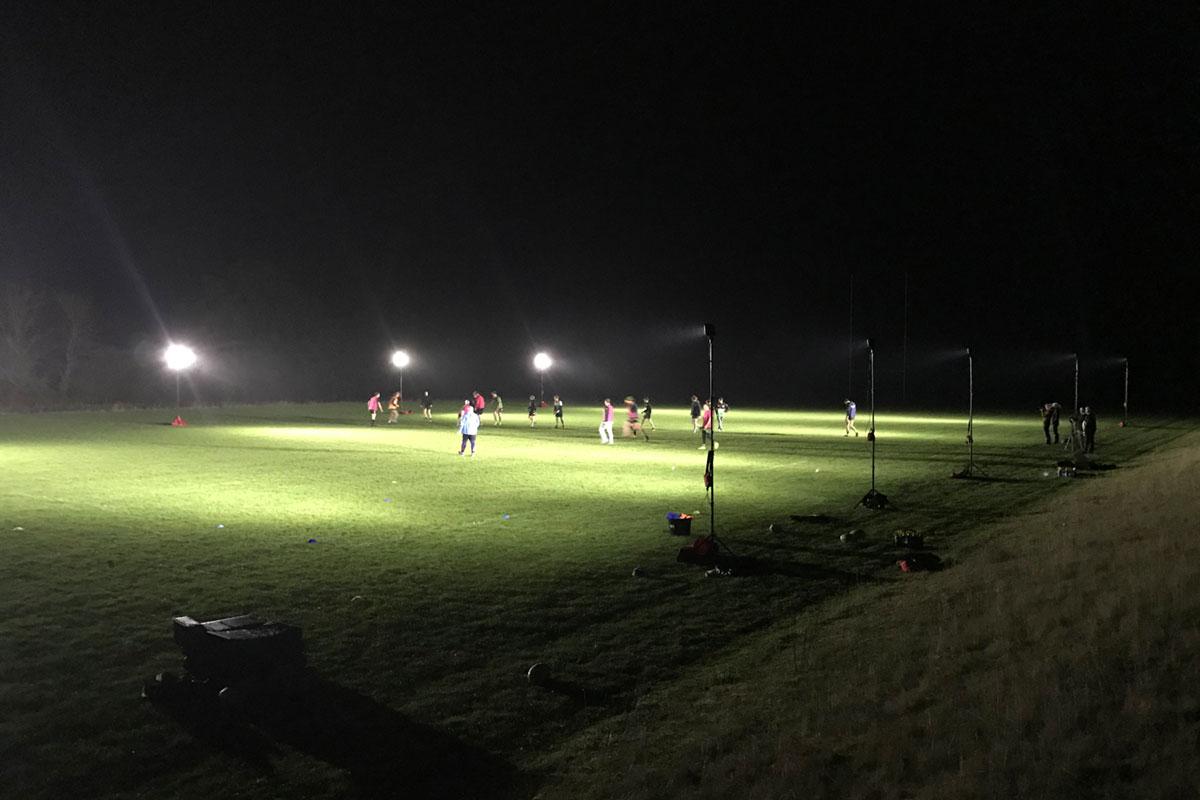 A group plays soccer on a grassy field at night, illuminated by the 20,000-lumen Nightsearcher SportStar Portable LED Floodlight, with equipment and shadows visible around the bright area.