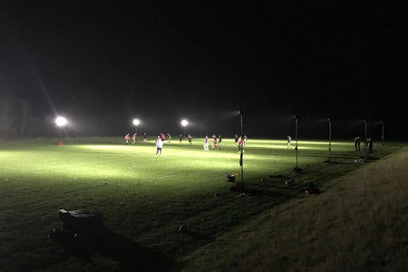 A group plays soccer on a grassy field at night, illuminated by the 20,000-lumen Nightsearcher SportStar Portable LED Floodlight, with equipment and shadows visible around the bright area.