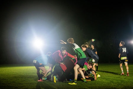 A group of rugby players compete for the ball at night on a grassy field, brightly lit by the Nightsearcher SportStar Portable LED Floodlight with 20,000 lumens in the background.