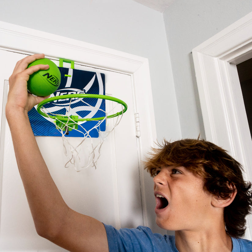 A boy in a blue shirt excitedly goes for a slam dunk with a green foam ball on the Nerf Nerfoop indoor basketball hoop, mounted over a white door.