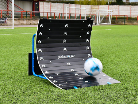 A soccer ball strikes the Precision Training PrecisionPLAY Skills Trainer Ramp Rebounder on green artificial turf, with a goalpost and fencing in the background.