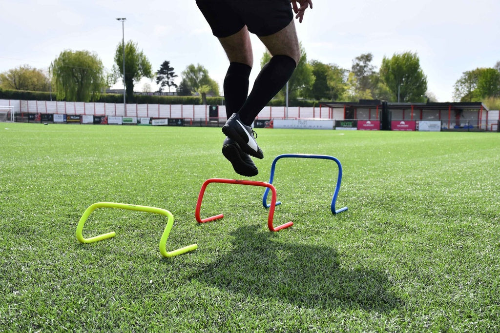 Wearing black sportswear, a person jumps over three Precision Training Precision Agility Hurdles—yellow, red, and blue—on a green soccer field, with a stadium and trees visible in the background.