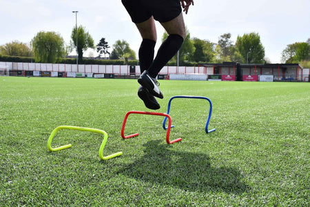 Wearing black sportswear, a person jumps over three Precision Training Precision Agility Hurdles—yellow, red, and blue—on a green soccer field, with a stadium and trees visible in the background.