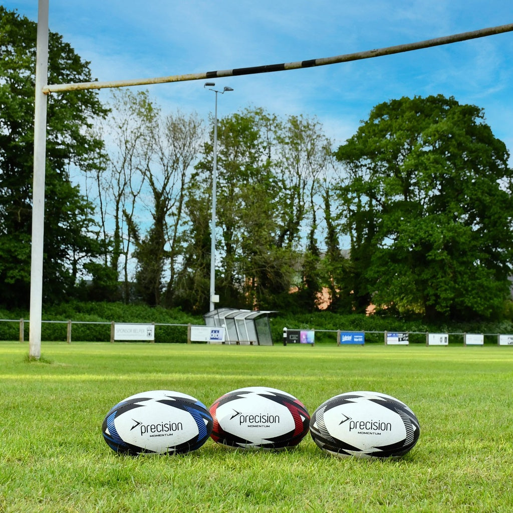 Three Precision Momentum Rugby Training Balls by Precision Training are arranged on a grassy field before a goalpost, offering reliable all-weather grip—perfect for rugby practice in any conditions.