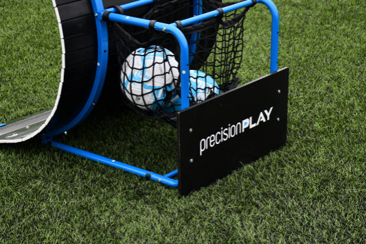 A close-up of the Precision Training PrecisionPLAY Skills Trainer Ramp Rebounder in blue and black, featuring two soccer balls in a net on green artificial turf, with a sign highlighting its use for football skills training.
