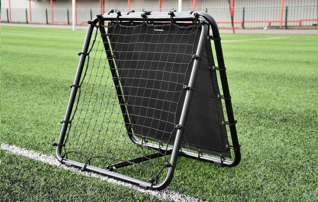 The Reydon Sports Precision Pro Double Sided Rebounder stands on green artificial turf near a white line, making it essential training pitch equipment. A metal fence and red posts are visible in the background.