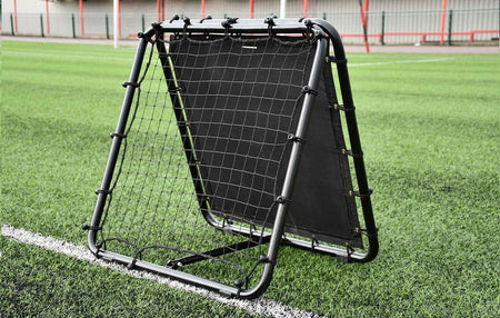 The Reydon Sports Precision Pro Double Sided Rebounder stands on green artificial turf near a white line, making it essential training pitch equipment. A metal fence and red posts are visible in the background.