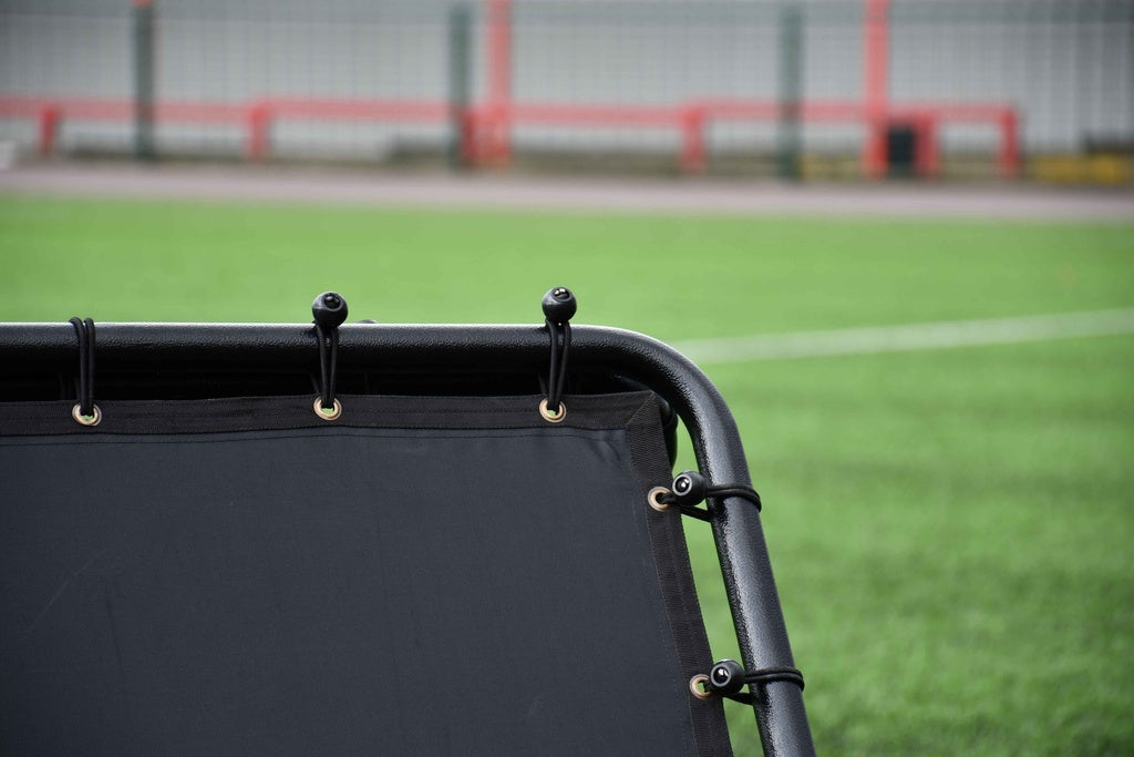 Close-up of the Precision Training Precision Pro Double Sided Rebounder, featuring eyelets and cords on green artificial turf, with blurred fence and red barriers—ideal soccer training net for enhancing skills.