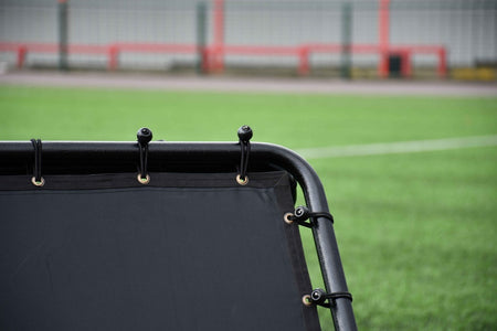 Close-up of the Precision Training Precision Pro Double Sided Rebounder, featuring eyelets and cords on green artificial turf, with blurred fence and red barriers—ideal soccer training net for enhancing skills.