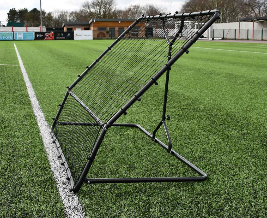The Precision Training Precision Pro Two Angled Rebounder, a key training tool, is set up on artificial turf beside a white sideline, with empty stands and trees visible in the background.