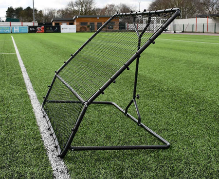 The Precision Training Precision Pro Two Angled Rebounder, a key training tool, is set up on artificial turf beside a white sideline, with empty stands and trees visible in the background.