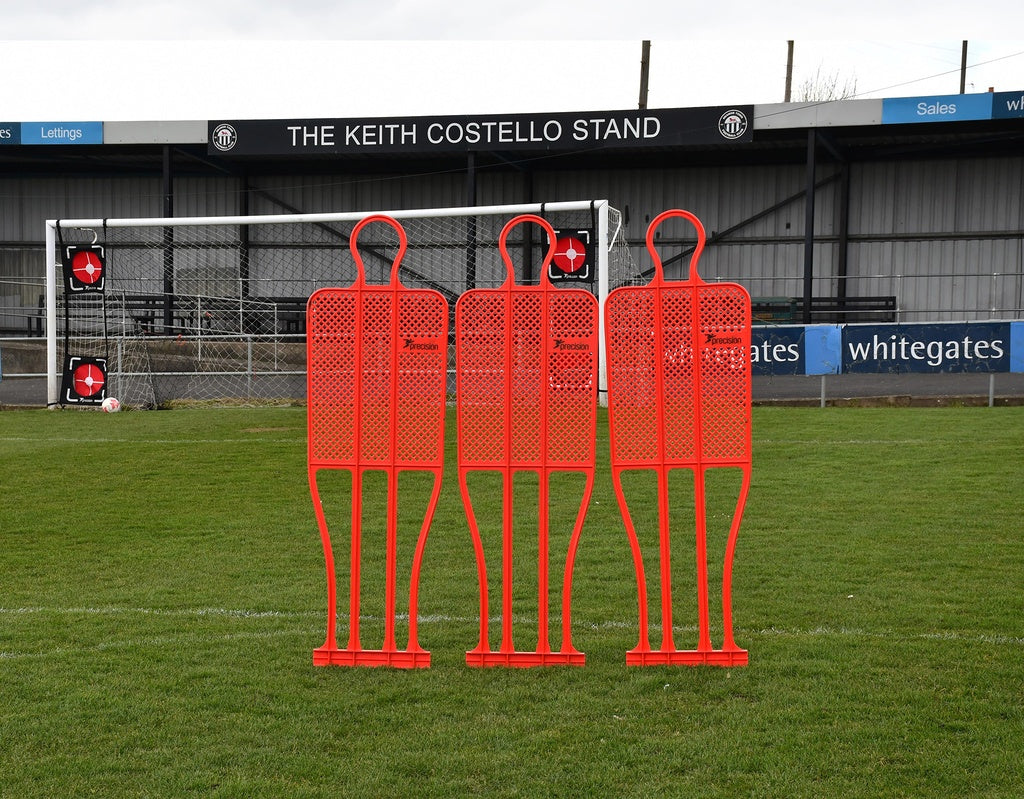 A Precision Training Precision Pro Club Mannequin (Single) in red stands on a grassy field by the goalpost—perfect for free kick drills—with empty stands and the sign "THE KEITH COSTELLO STAND" visible in the background.