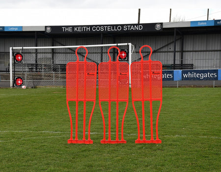 A Precision Training Precision Pro Club Mannequin (Single) in red stands on a grassy field by the goalpost—perfect for free kick drills—with empty stands and the sign "THE KEITH COSTELLO STAND" visible in the background.