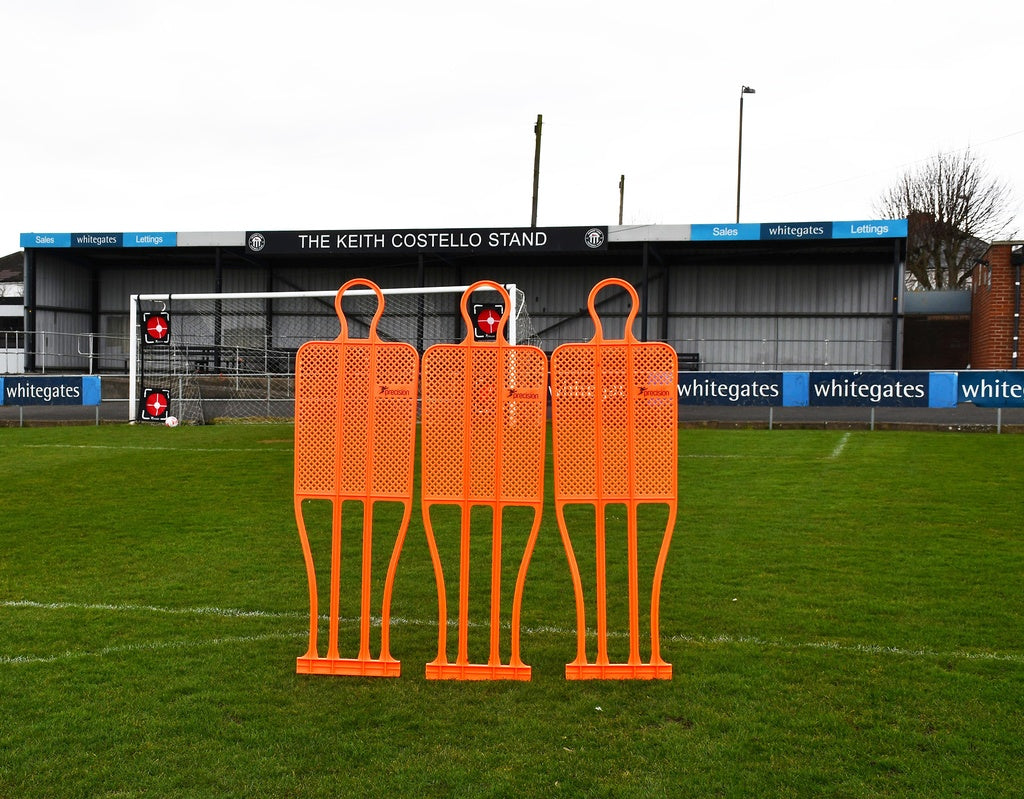 A Precision Pro Club Mannequin (Single) by Precision Training in fluo orange stands on a soccer field near the goal, set up for free kick practice, with empty stands and advertising boards behind.