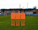 A Precision Pro Club Mannequin (Single) by Precision Training in fluo orange stands on a soccer field near the goal, set up for free kick practice, with empty stands and advertising boards behind.