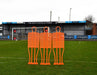 A Precision Pro Club Mannequin (Single) by Precision Training in fluo orange stands on a soccer field near the goal, set up for free kick practice, with empty stands and advertising boards behind.