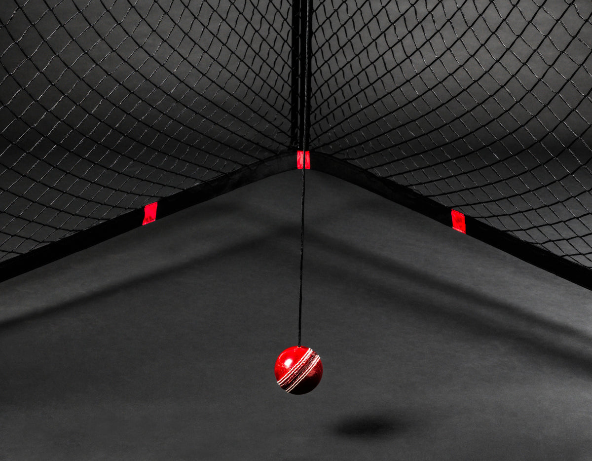 The V Pro 2.0 by The V features a red cricket ball suspended on black netting, with dramatic lighting and shadows creating an intense cricket training ambiance against a dark background.