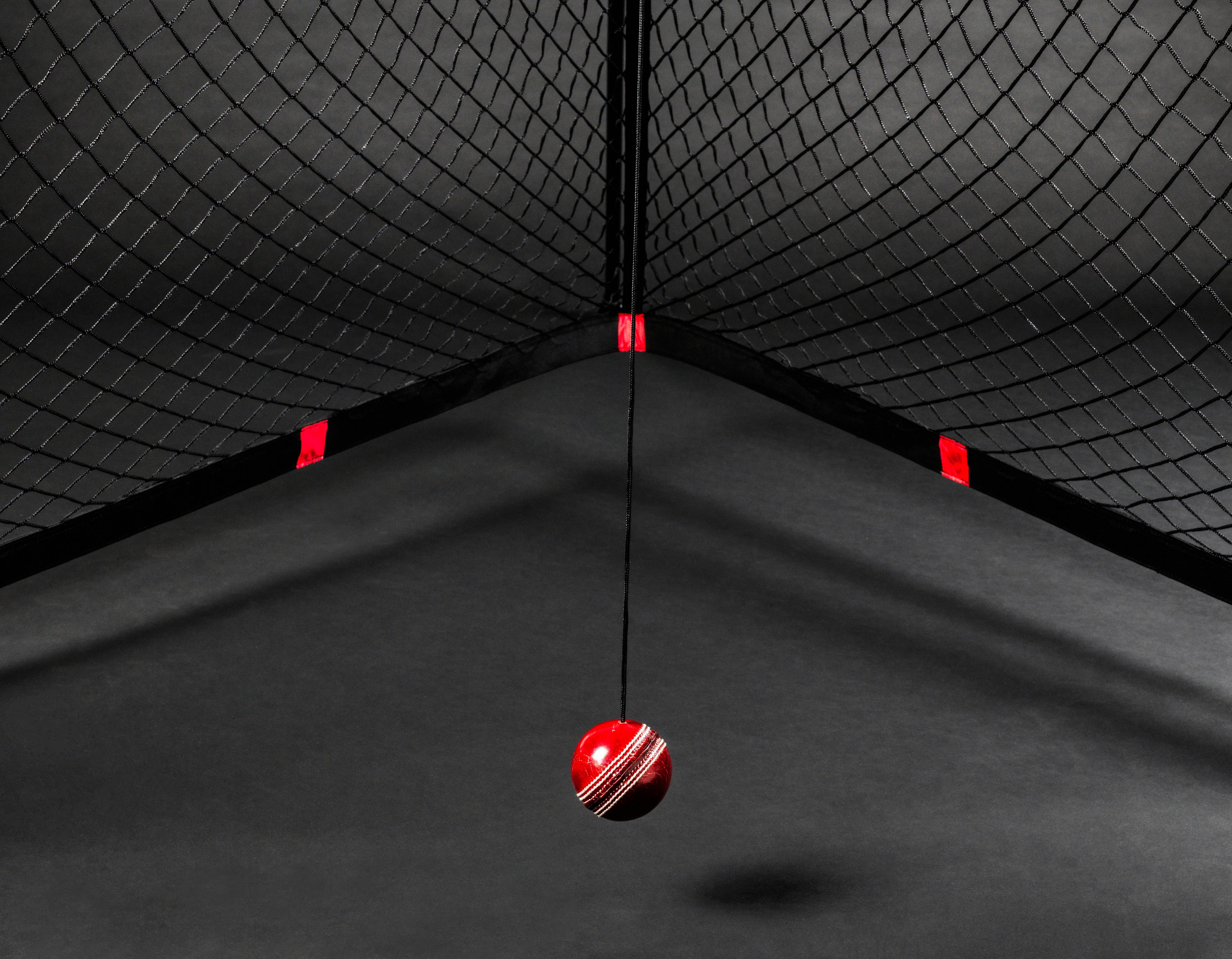 The V Pro 2.0 by The V features a red cricket ball suspended on black netting, with dramatic lighting and shadows creating an intense cricket training ambiance against a dark background.
