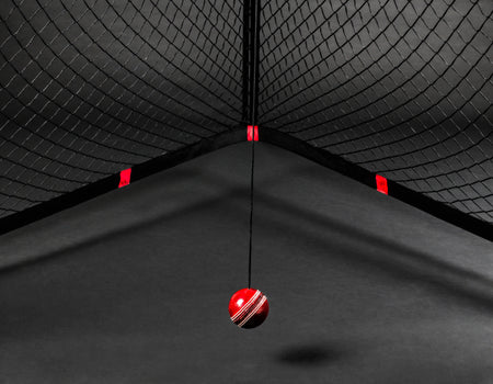 The V Pro 2.0 by The V features a red cricket ball suspended on black netting, with dramatic lighting and shadows creating an intense cricket training ambiance against a dark background.