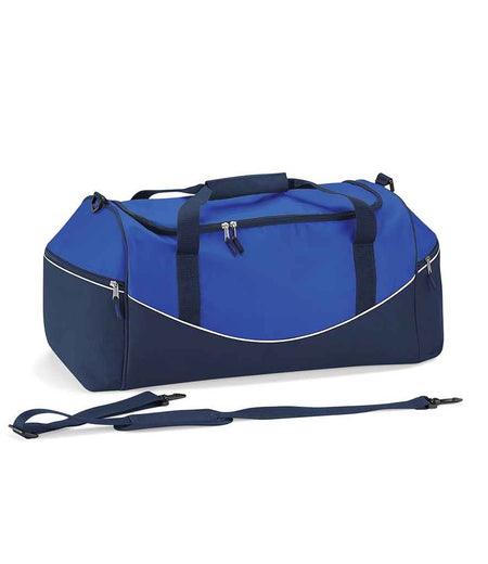 The Quadra Teamwear Holdall by Quadra features a royal blue/navy/white design, black handles, zipper pockets, white piping accents, padded hand grip, large end pockets, and a detachable shoulder strap in front of the bag.