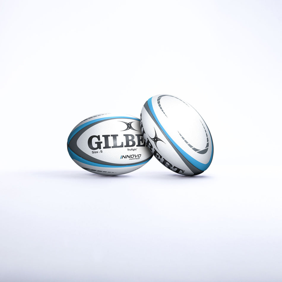 Two Gilbert Innovo Match Balls (Size 5) by Gilbert, white and blue, are displayed on a plain white background—one upright and one lying on its side with the logo clearly visible.