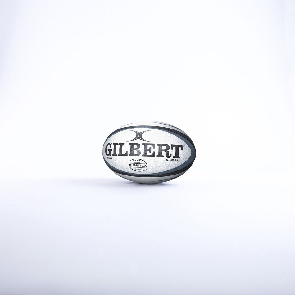 A Gilbert Kinetica Match Rugby Ball Size 5 by Gilbert, featuring black and gray markings, rests on a white surface against a plain white background.
