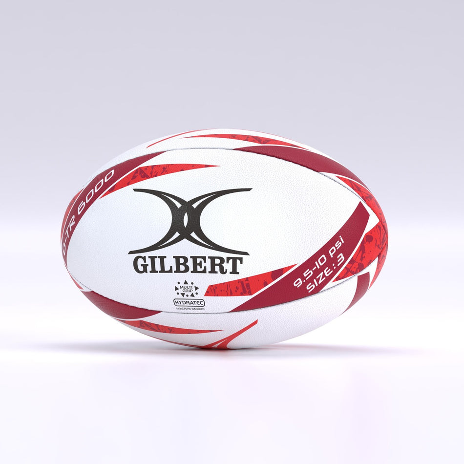 The Gilbert G-TR6000 Training Rugby Ball by Gilbert features a white and red design with black branding, a durable rubber surface, and rests on a smooth surface—ideal for schools and clubs.