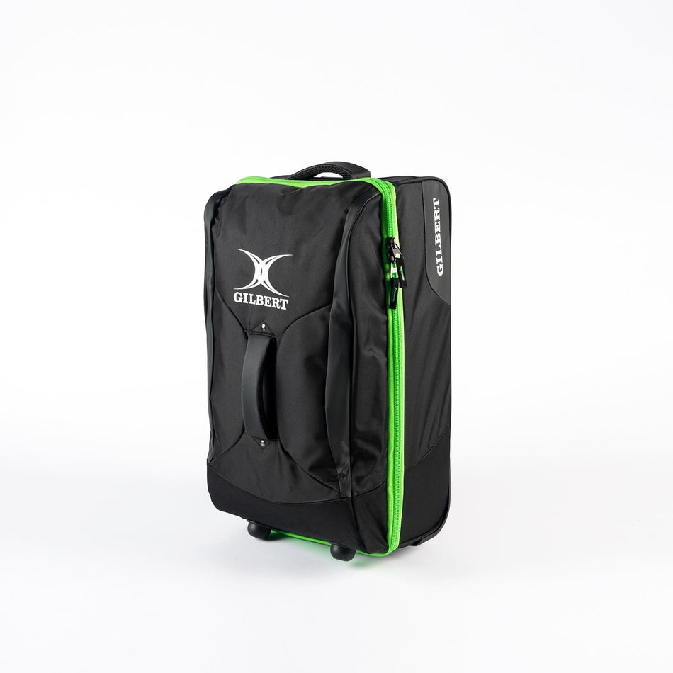 A black Gilbert Flight Bag stands upright on two wheels against a white background. It features sectioned storage, top and side handles, a green zipper, and the Gilbert logo in white on the front.