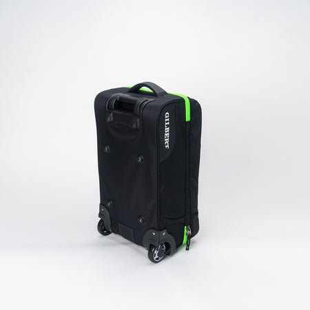 The Gilbert Flight Bag, by Gilbert, is black with green zippers, two wheels, sectioned storage, a top handle, and branding on the side. It is displayed against a plain white background.
