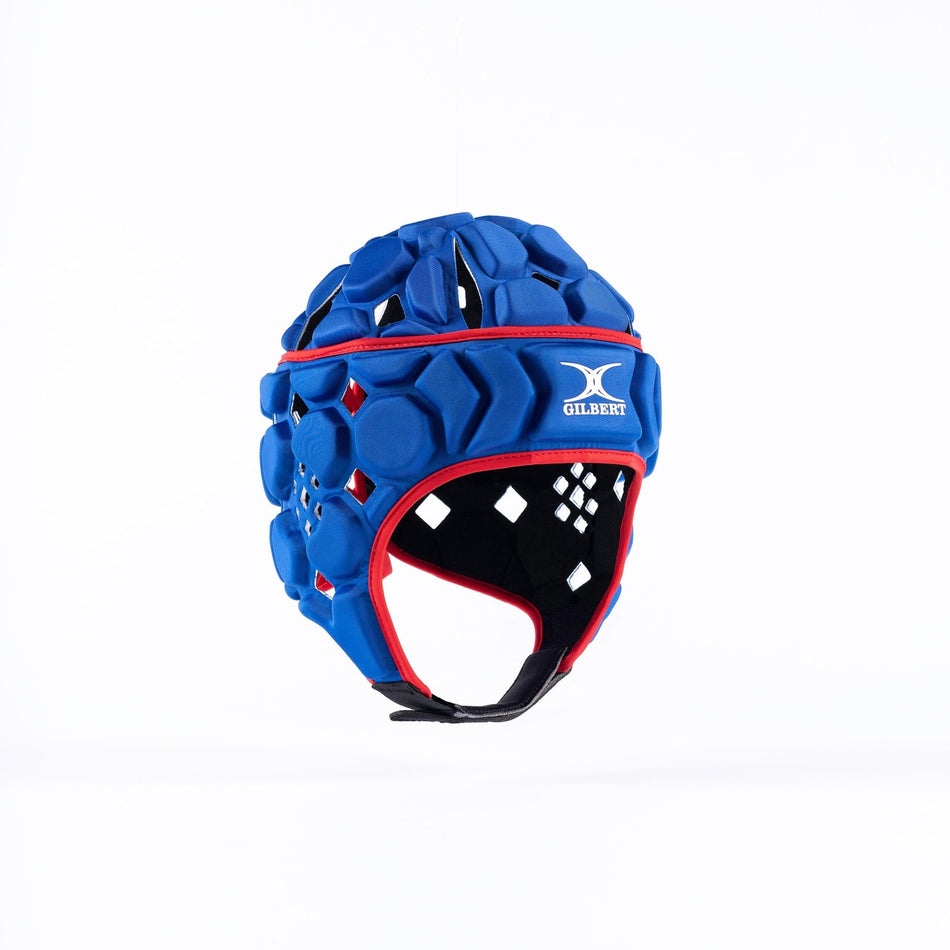 The Gilbert Octane Senior Headguard features royal blue hexagonal padding, red trim, black chin strap, a white Gilbert logo on the front, and an expandable lace closure for a secure fit. Shown on a plain white background.