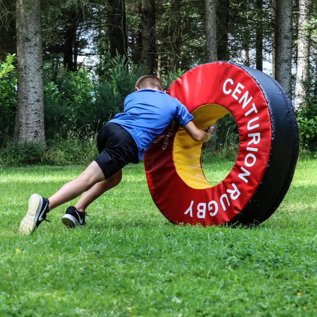 A boy in a blue shirt and black shorts pushes a large red and yellow Centurion Foam Tackle Ring (Doughnut), a rugby training aid, across a grassy field with trees in the background.