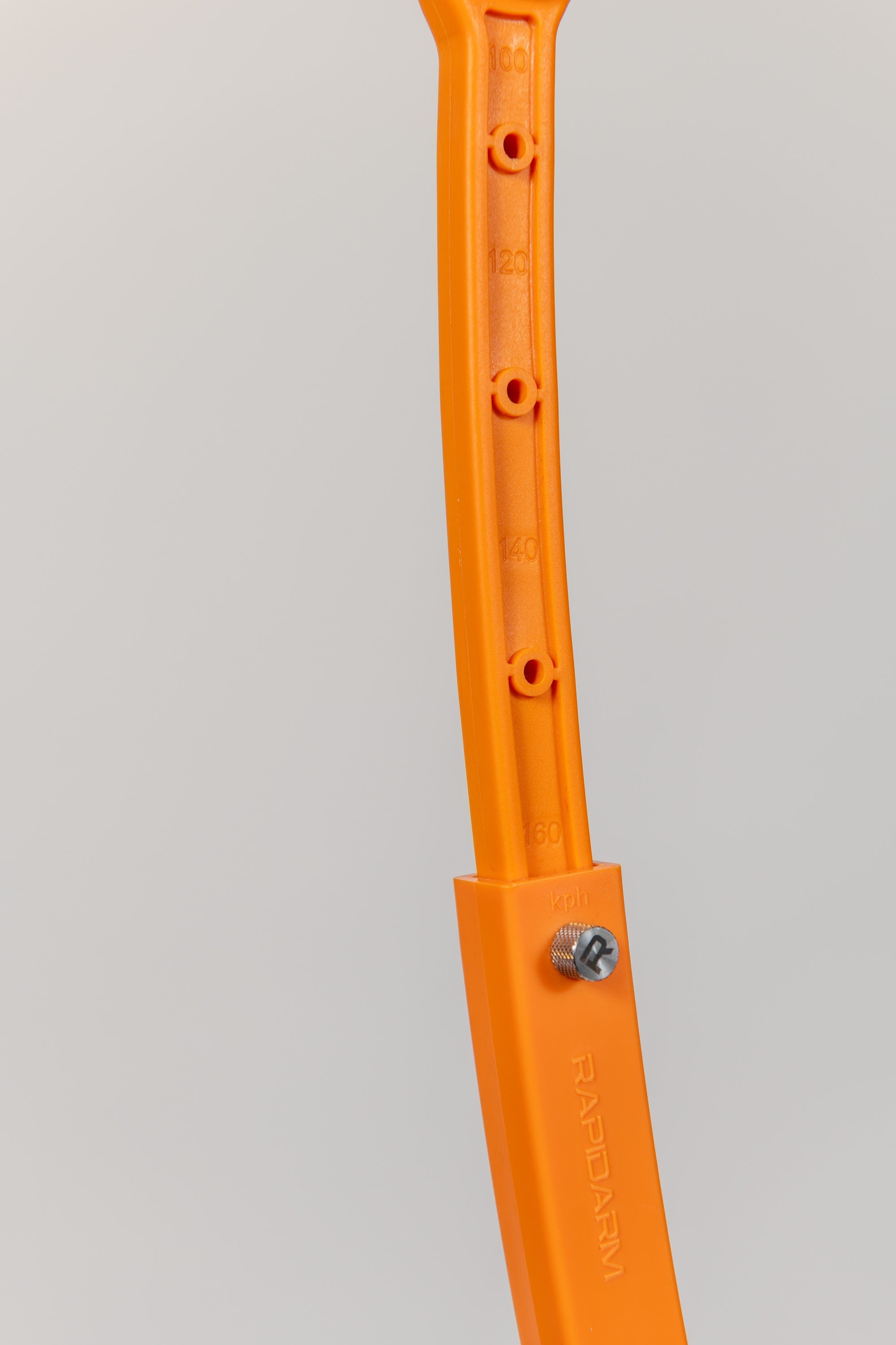 Close-up of a RapidArm+ Ball Thrower by RapidArm, featuring orange plastic with measurement markings, holes, a screw, and the embossed brand name. The adjustable cricket training arm is set against a light plain background.