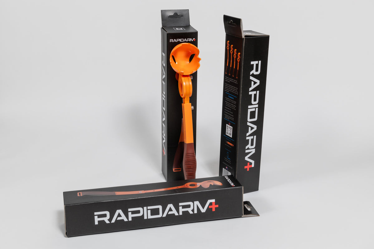 A vibrant orange RapidArm+ Ball Thrower by RapidArm, featuring an adjustable arm, is displayed both inside and outside its black packaging, alongside three boxes set against a plain white background.