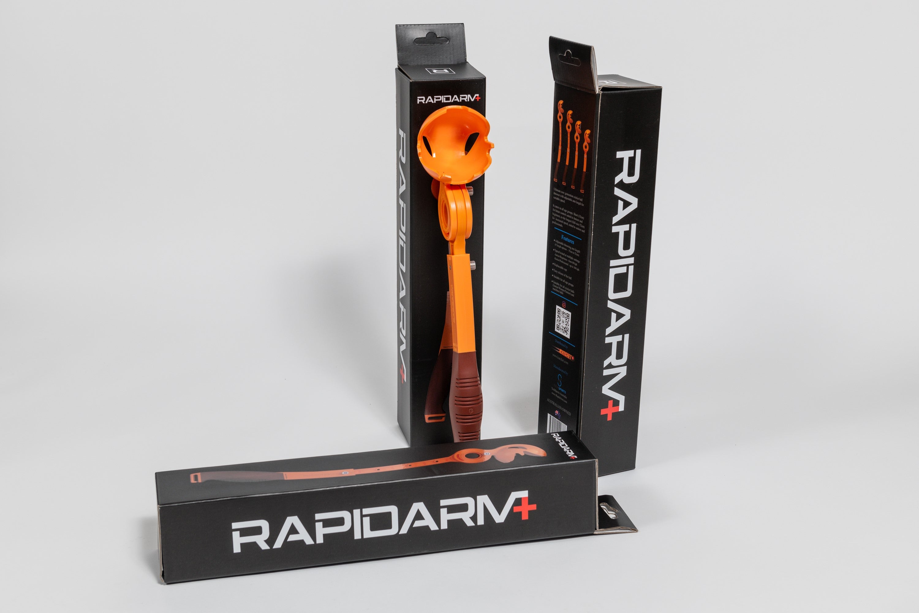 A vibrant orange RapidArm+ Ball Thrower by RapidArm, featuring an adjustable arm, is displayed both inside and outside its black packaging, alongside three boxes set against a plain white background.
