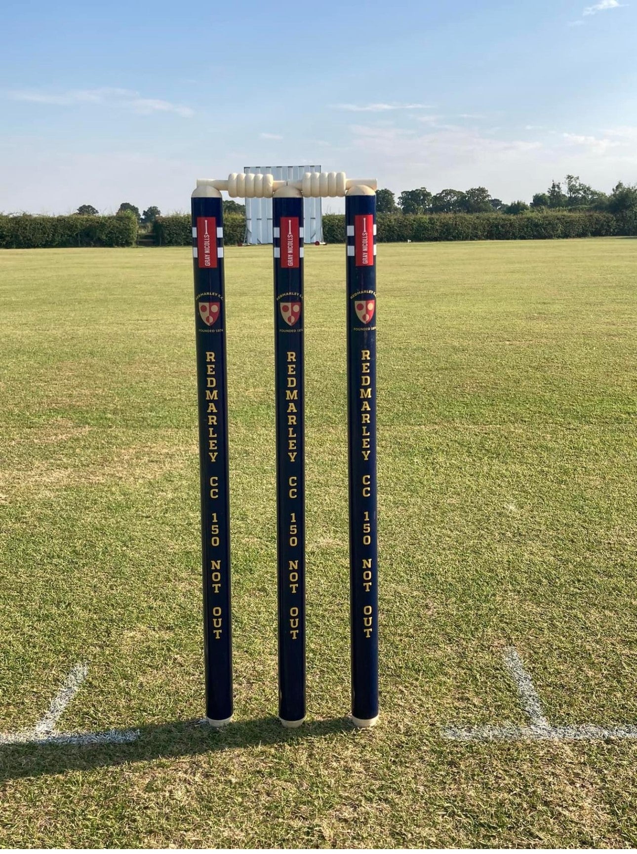 Three Custom Printed Club Cricket Stumps by Gray Nicolls, personalised with "REDMARLEY CC 150 NOT OUT," stand on a grass field with white bails, set against a backdrop of hedge, trees, and clear sky.