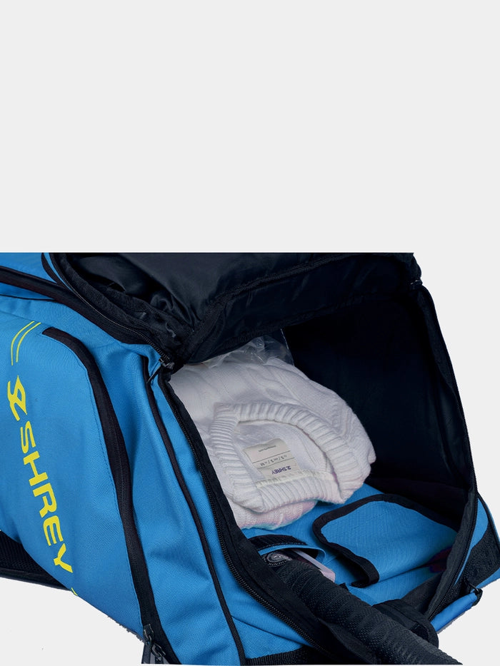 A blue Shrey Legacy Duffle Wheelie 80 cricket kit bag with yellow "SHREY" branding is unzipped, showing neatly folded white clothing inside.